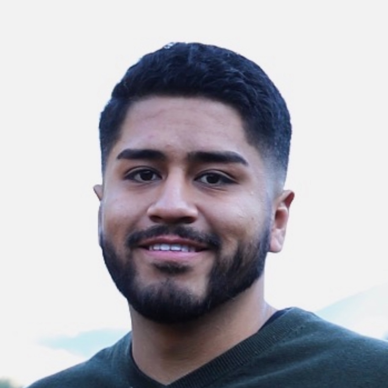 Erick Alatorre, a man with black hair, brows, and facial hair, grins, with a white background.