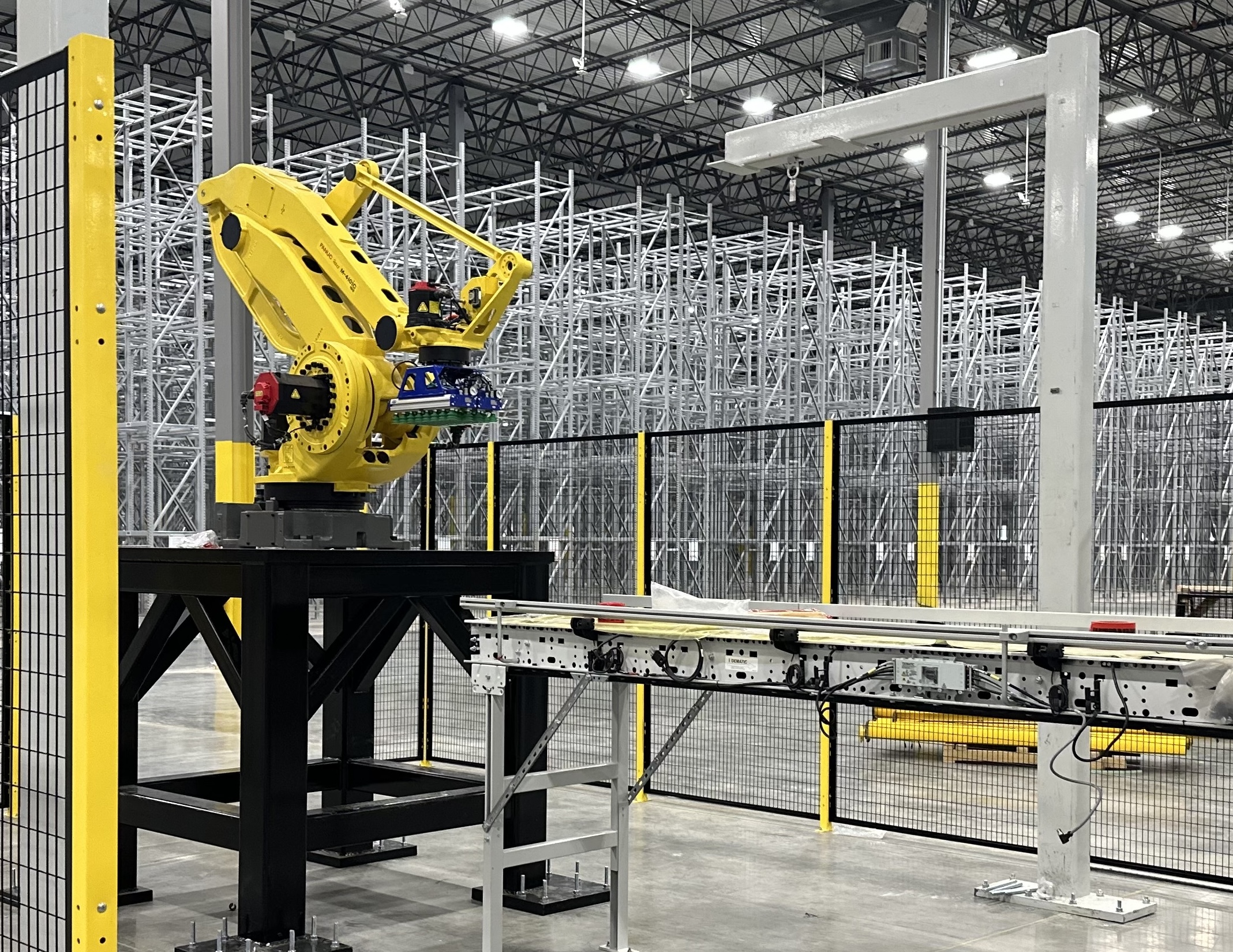 A white AS/RS in the background and a yellow articulated robot arm in the foreground.