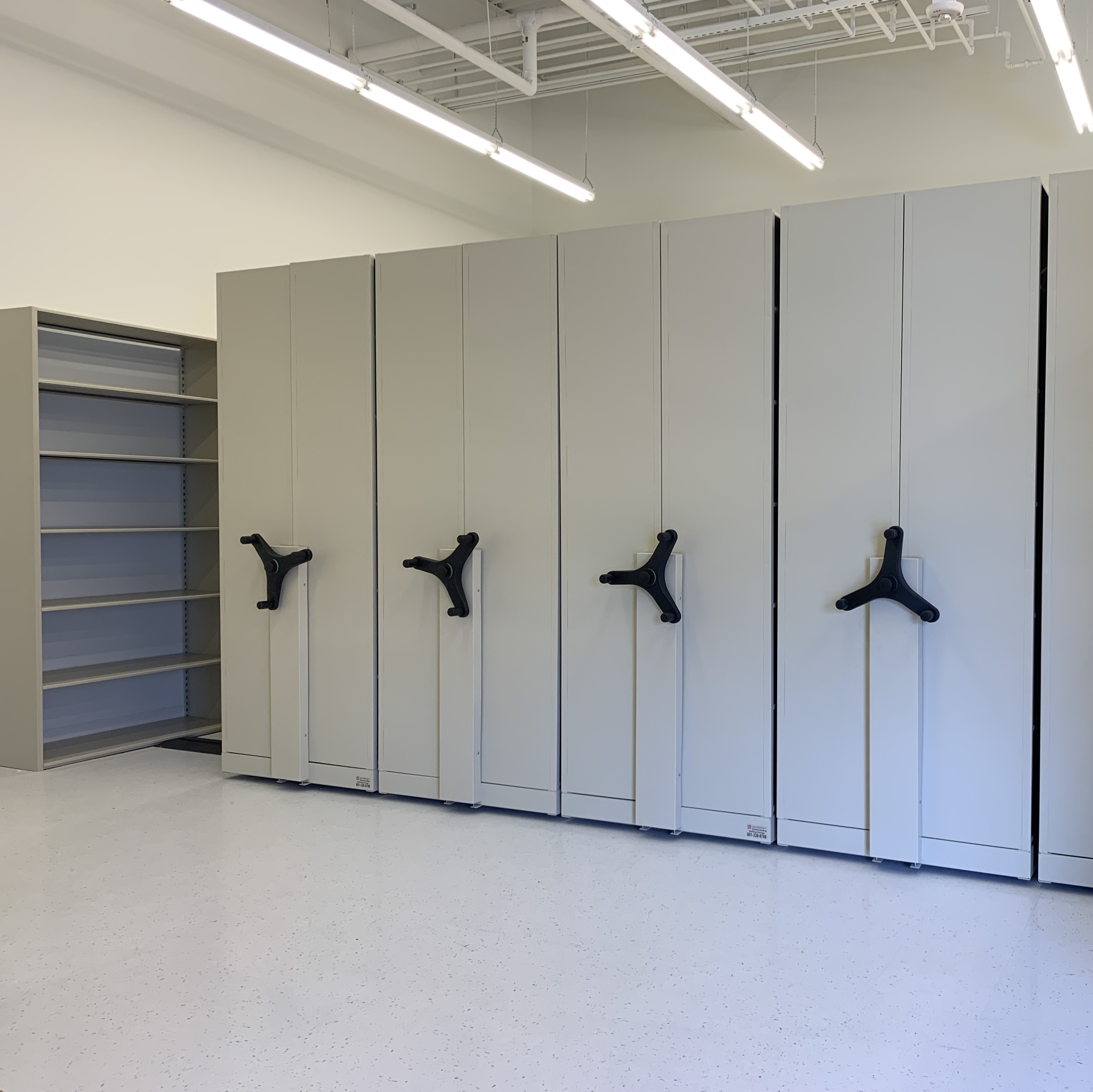 Shelves on tracks, each with cranks on the sides; most shelves are pushed against one another except for a single aisle.