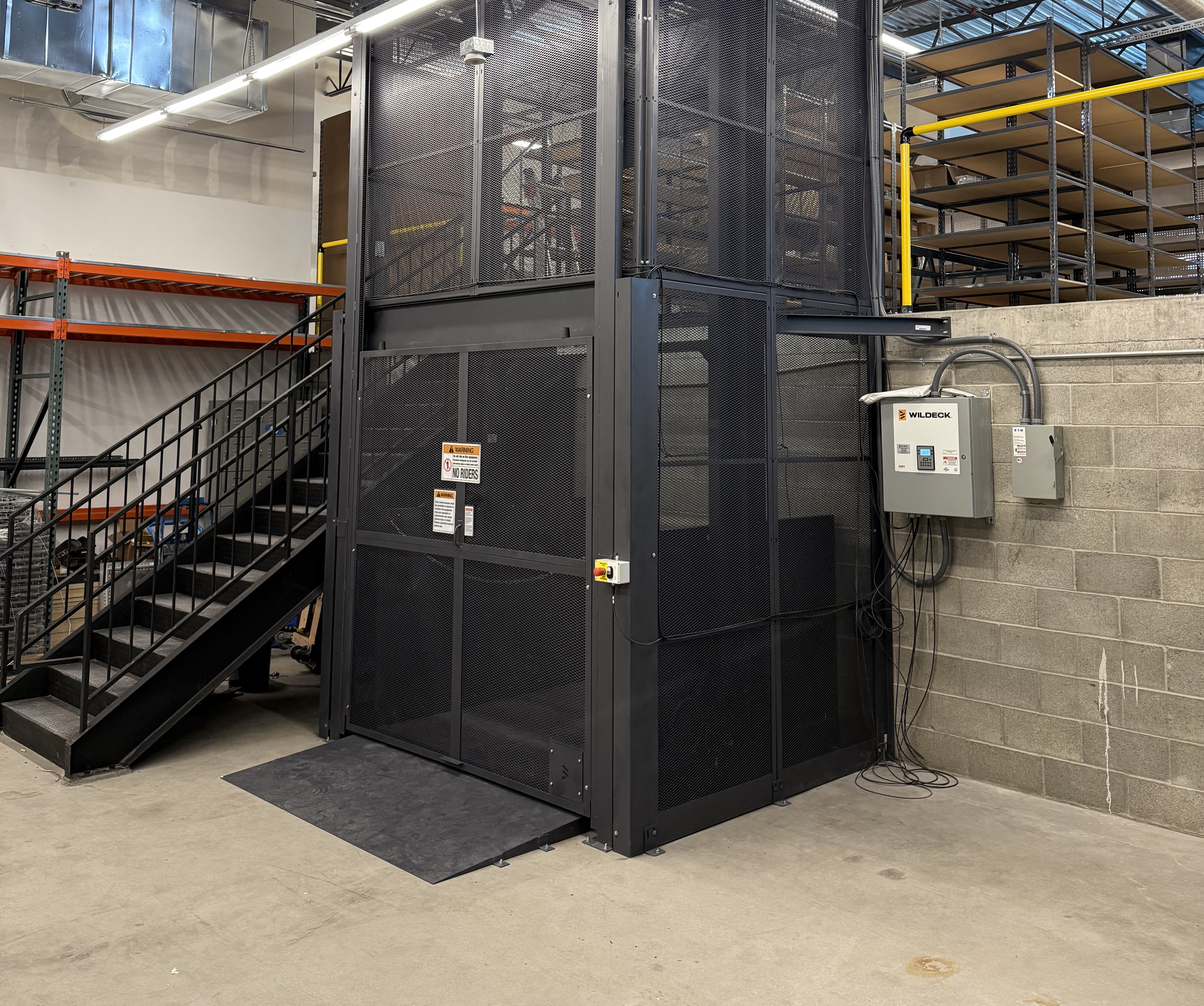 A black freight lift elevator against a large ledge and next to a staircase.